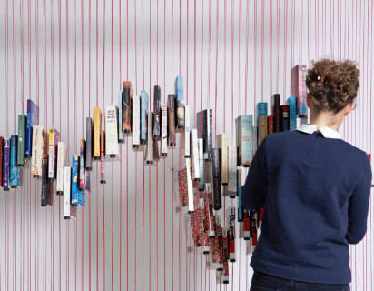 Paperback books suspended on vertical red strings at different heights, creating a zig-zag pattern. To the right of the image, a woman stands with her back to the camera, adding or removing books from the string ‘bookcase’