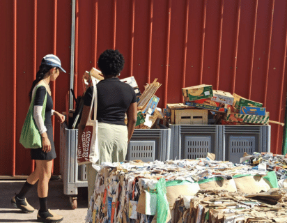 Two women stand with their backs to the camera, looking at a recycling unit which is overflowing with wooden crates and cardboard boxes. Cubes of compacted materials for recycling stand in the foreground, slightly out of focus