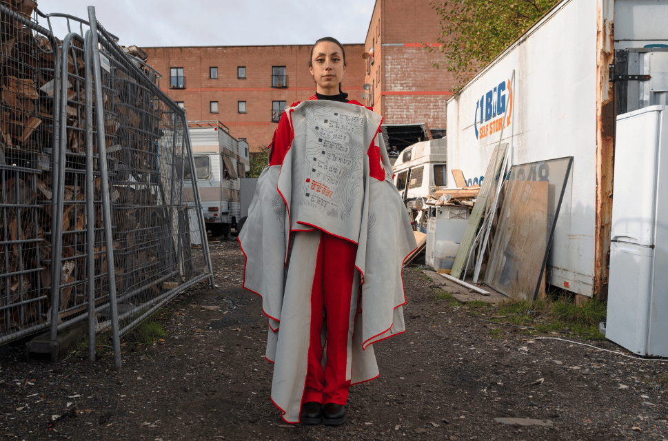 Woman stands in scrap yard with buildings behind and broken fencing to her left. She is wearing an outfit of red and white and has her hair tied back.