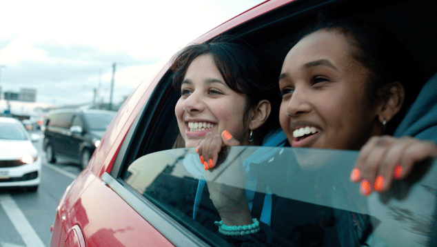 A still from 'Brides', directed by Nadia Fall. The still shows two young women looking out of the open window of a moving car on a motorway. Both are smiling.