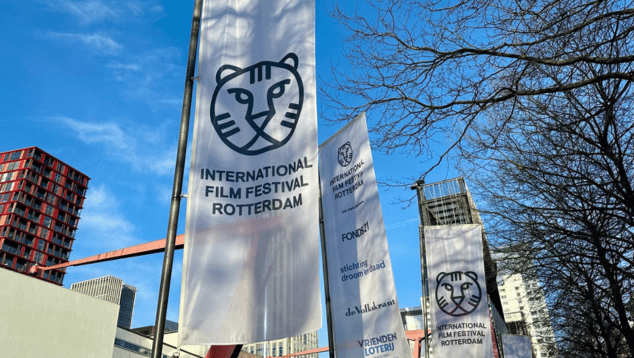 Banners for the Rotterdam Film Festival photographed from underneath against an urban skyline with bright blue sky and a tree with bare branches on the right of the image.