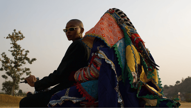 A still from 'Egungun' directed by Olive Nwosu. A woman with a bald head wearing sunglasses rides a motorbike with a figure sitting on the back, shrouded in colourful blankets.