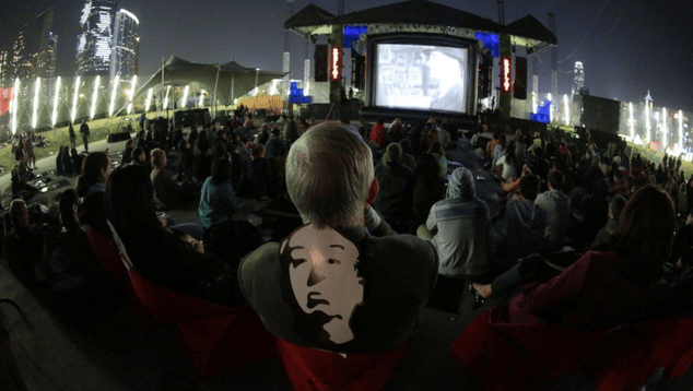 A film festival-goer photographed from behind watches an outdoor film screening amongst a crowd. Around their neck is a paper mask of the face of filmmaker Alfred Hitchcock.