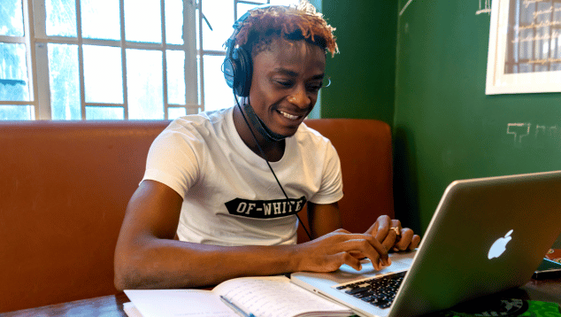 A young man works at a laptop with an open notepad to his right. He is smiling and wearing headphones, apparently engaged in an online call.