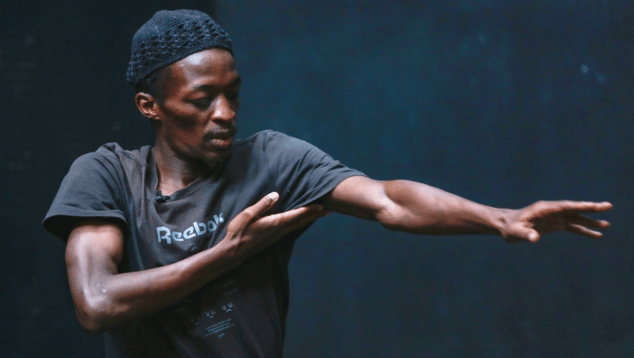 A male performer, Kaldi Makutike, stands with his left arm outstretched, and his right hand reaching towards his left arm. Photo by Zivanai Matangi (c) British Council