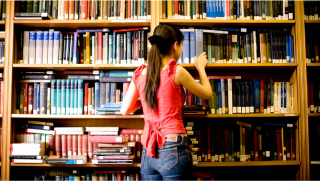 A young woman with long dark hair stands with her back to the camera, taking a book out of wall-mounted bookshelves.