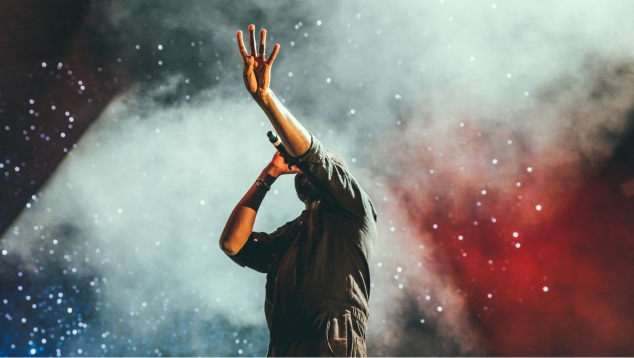 Man performing with bright lights behind him, holding a microphone in one hand and four fingers up in the sky with the other.