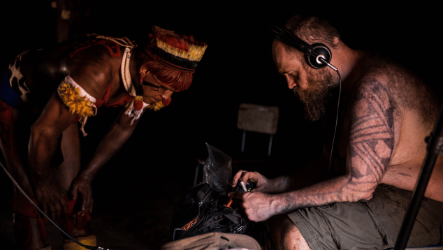 Two men either side of a device one is holding between them while also wearing headphones. Musicians in Residence Brazil Nathaniel Mann Wauja. Photo by Jean Nunes (c) British Council