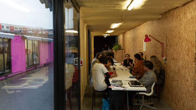 A group of people sit around a table in an industrial style cabin under a strip light.