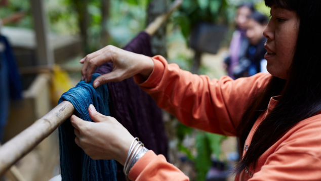 A woman hangs freshly dyed fabric over a pole suspended between trees in a woodland.