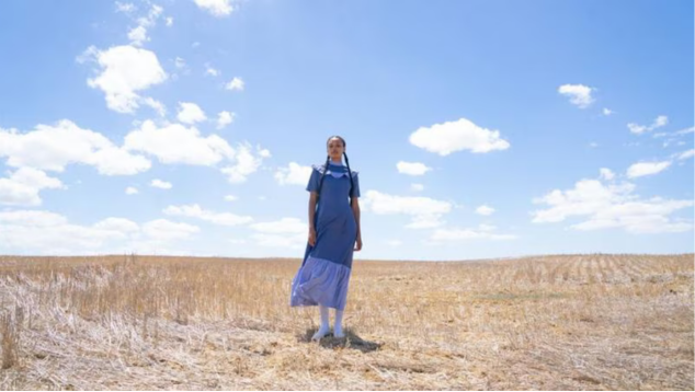 Desginer Sindiso Khumalo, AW2021, South Africa © Sindiso Khumalo. A woman wearing blue with her hair tied in pigtails stands alone in a barren meadow against a blue sky with white clouds.