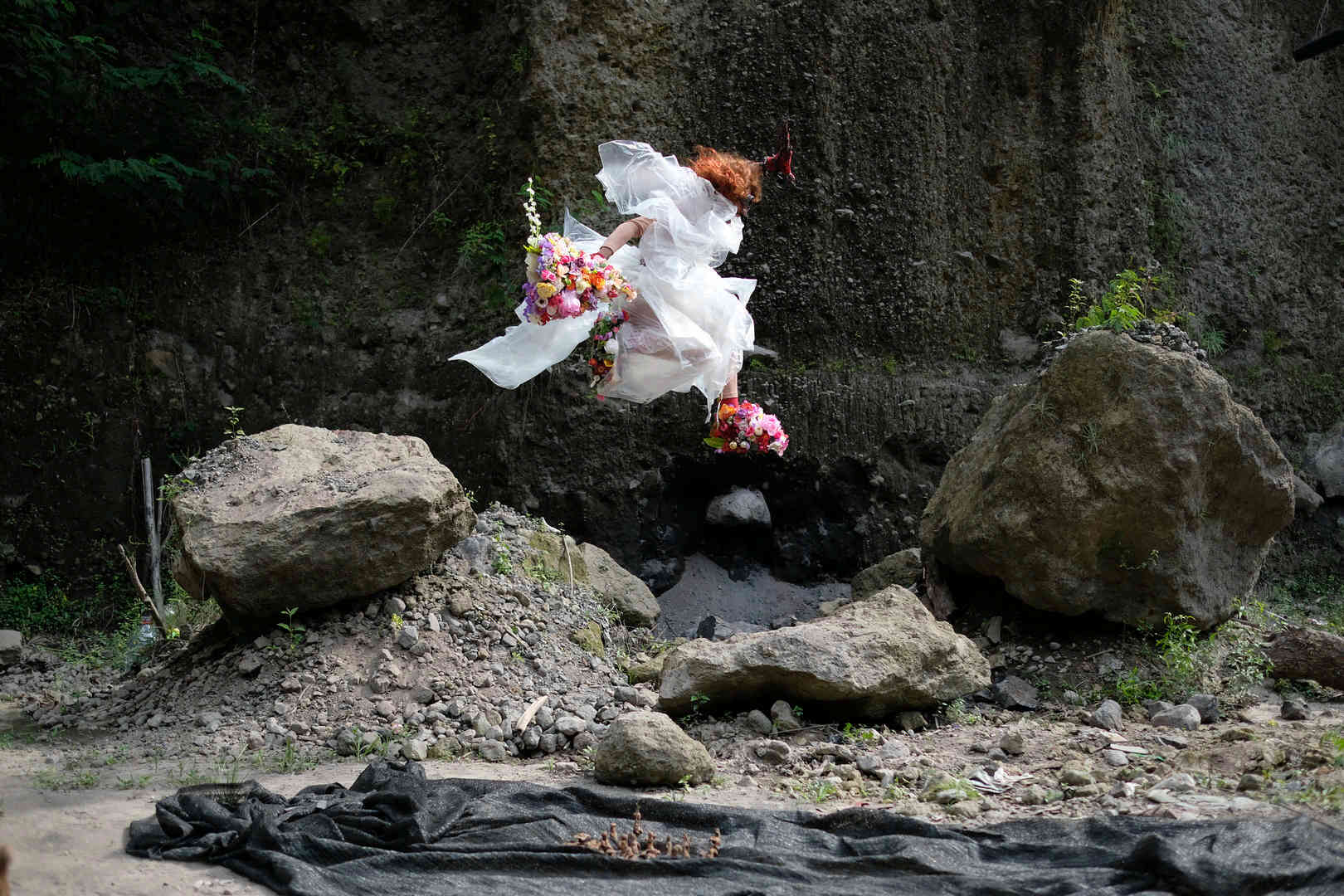 A woman wearing a white dress with a bouquet of flowers in her hand and flowers on her feet leaps over rocks next to what looks like a cliff face. Photo by Sito Adi Anom & Ariyanto Nugroho