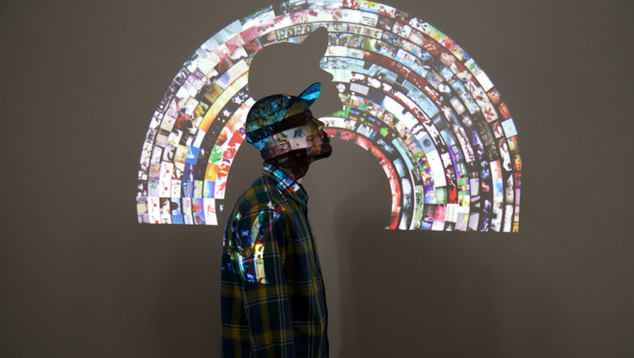 A man wearing a baseball cap stands in profile against a wall with a projected image of mosaic in the shape of a rainbow.