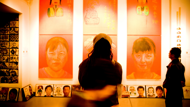 Two people stand in a gallery space, silhouetted. They are looking at a wall displaying three large art works of portraits of women.