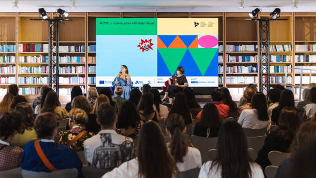 In conversation with Katy Hessel at WOW Athens 2024. An audience watches an event with two seated speakers sitting in front of a screen displaying branding for WOW - Women of the World. The event appears to be taking place in a library. Photo by Euftychia Vlachou