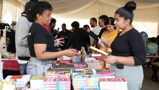 Many people in a marquee with two people at the front of the picture talking to each other, one holding books, next to books.