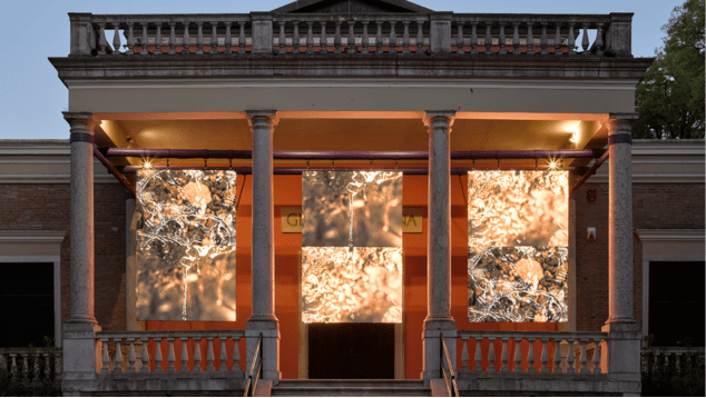 The portico of the British Pavilion building in Venice at dusk. The portico is lit from the rear, with three large screens hanging in the spaces between four pillars. The screens show close ups at different angles of water with sunlight reflecting off its surface. Photo by Jack Hems (c) British Council