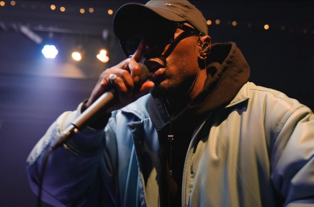 Close up of man, holding microphone to his mouth, wearing hooded top, cap and sunglasses. Bright indoor stage lights behind him.