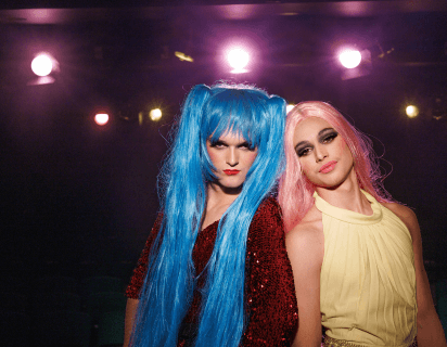 Two teenage boys dressed in drag stand side by side, shoulder to shoulder to the right of the frame. The boy on the left wears a bright blue wig in pigtails and the boy on the right wears a long pink wig. There are bright lights, resembling stage lighting, above their heads and they stand against a dark background.