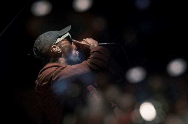 Man wearing cap and sunglasses, holding mic, with a dark background and blurred lights around him.