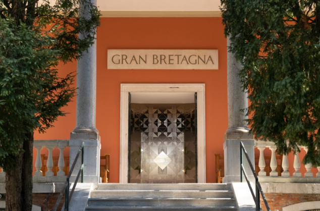 Outside, looking towards a large open door at the top of several steps with 'Gran Bretagna' sign above it. Two trees at the side of the steps.