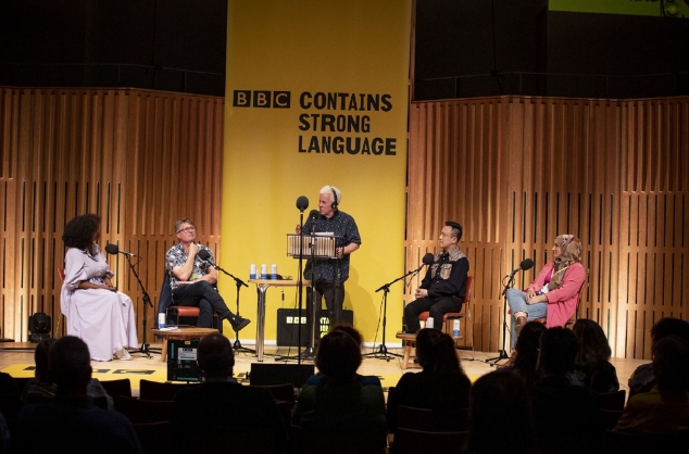 A stage featuring five people, the one in the centre is stood up, the others are sat down. Behind him, a yellow poster says BBC Contains Strong Language.