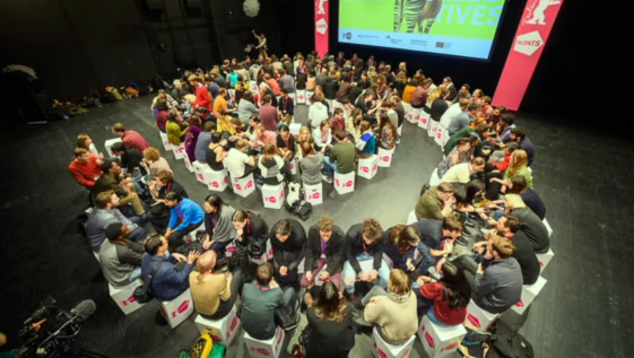 Many people sat opposite each other on small stools in front of projection screen.