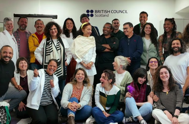 A group of people, some stood, some sitting on the floor in an office, all smiling, some looking at each other.
