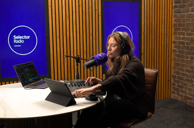 Lady in radio studio with headphones on, microphone in front of her, at a desk, two laptops next to her, Selector Radio logo on a screen behind her.