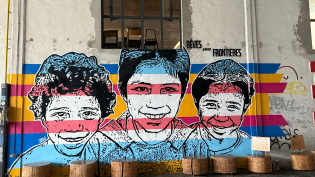 A mural of three children's smiling faces, with stripes of yellow, blue, white and red painted across them. The mural is painted on a wall painted white with a window above through which three chairs are visible. Friche La Belle Mai, Marseille (2025) Credit Marta Foresti