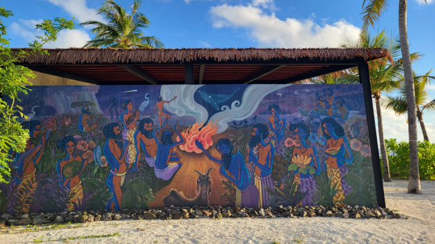 A mural painted on a building/shelter with a thatched roof on a beach in the Maldives. The mural shows people engaged in a ceremony around a fire. Image by Genevieve Pace. Oaga Art Resort collaborates with Maldivian artists and craftsmen, ensuring the resort features the largest art collection in the Maldives. The artwork is inspired by Maldivian folklore, mythology, and daily life, offering guests an immersive experience into the island’s rich heritage.