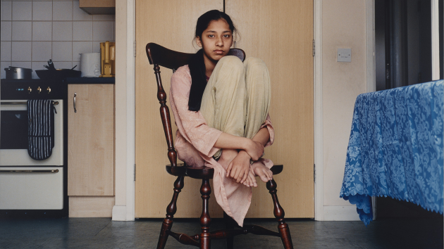 A woman sits on a wooden chair in the middle of a kitchen, with her knees bent and feet placed on the seat in front of her, and her hands clasped around the front of her ankles. Suki Dhanda, Untitled, C-type mounted on aluminium, 2002. Courtesy of the artist