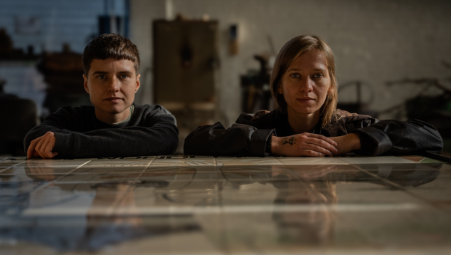 Two people sit alongside each other with their arms resting on a shiny tabletop. They are looking straight ahead, into the camera.
