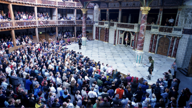 Two performers stand on the stage of Shakespeare's Globe Theatre in London, watched by a standing audience. Image (c) Getty