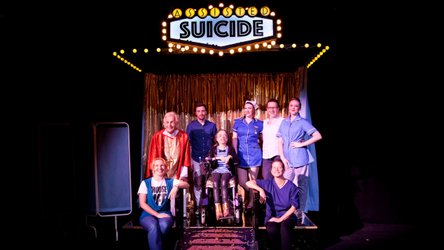 Liz Carr, Assisted Suicide the Musical at Southbank Centre Unlimited Festival. Photo by Rachel Cherry   A group photo of Liz Carr and her ensemble on stage. Above the group is a sign lit with lightbulbs as if it’s the outside of a theatre or cinema. On the sign is the title of the show ‘Assisted Suicide’. 