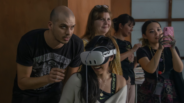 Indoors, one woman wearing a VR headset smiles while a man behind her guides her, looking above her. Three other women are behind them –  one is taking a photo.