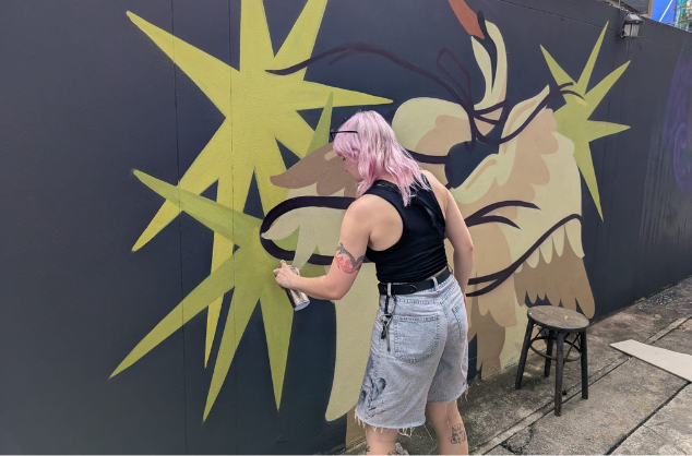 A woman paints a mural of abstract star-like shapes in yellows and greys. Belfast artist Zippy at work in Bangkok © Credit: Hit the North