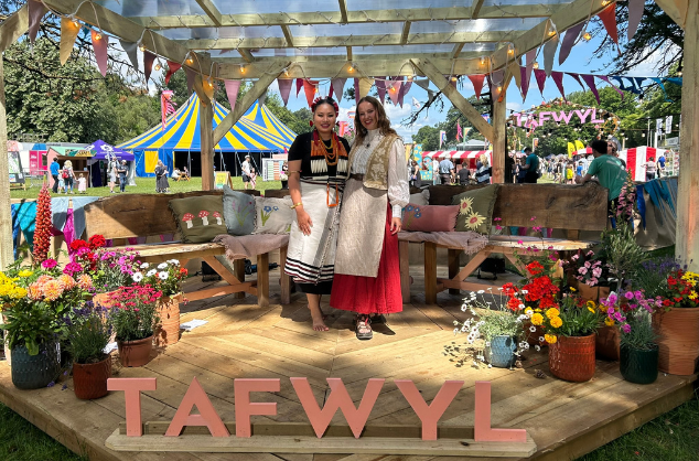 Two women stand on an outdoor platform/stage surrounded by flowers and bunting, wearing traditional Welsh dress. Mari and Seyie at Tafwyl 2025 (c) British Council