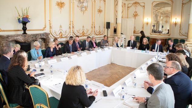 Large, formal room with many people sat around the table.