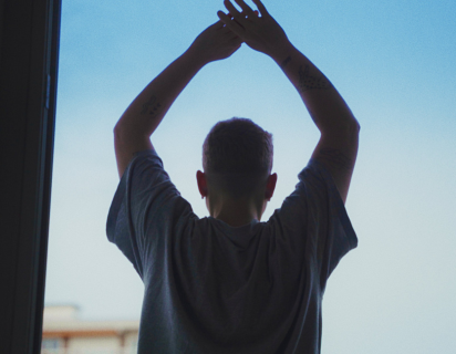 Still from 'Room 206' directed by Laurie Bisceglia (2025), part of Five Films For Freedom 2026. A young man stands in an open doorway, facing away from the viewer, silhouetted against a pale blue sky with his arms held up above his head, and his hands joined together.