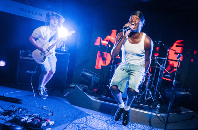On stage, electric guitarist on left, jumping into the air; vocalist on right wearing white vest also jumping.