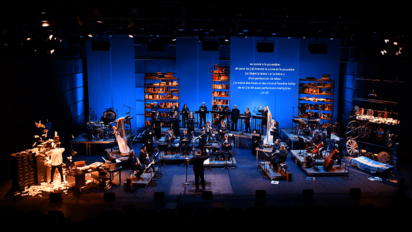 Orchestra performing on stage with blue lighting and a large screen displaying text behind them.