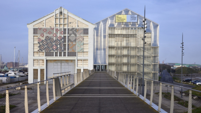 Wooden pedestrian bridge leading to two modern buildings with glass and patterned facades.