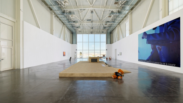 Spacious modern art gallery with high ceilings, large windows, and minimalist installations including a teddy bear on the floor.