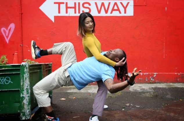 A dancer supports another dancer against a red wall with a sign saying 'this way'