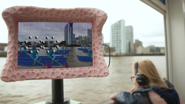 Digital screen on a boat showing animated fish over a city. On the boat, behind the sceen, there's a passenger wearing headphones looking out to  the river.