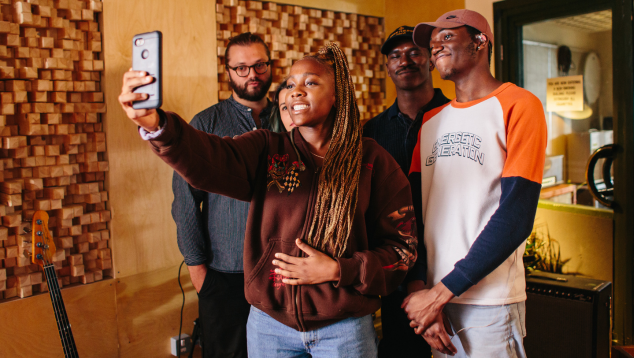 Five people in a recording studio, stood with the young lady at the front holding a camera to take a picture of them all.
