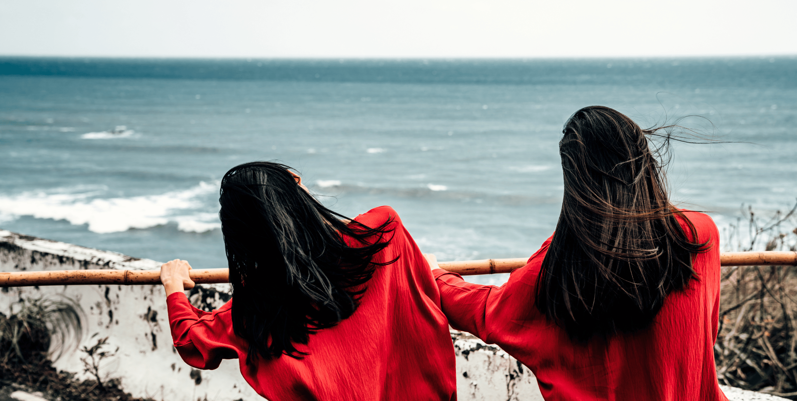 Two girls with long dark hair wearing red stand in the foreground by a seafront, looking out to see holding onto a rusty rail.