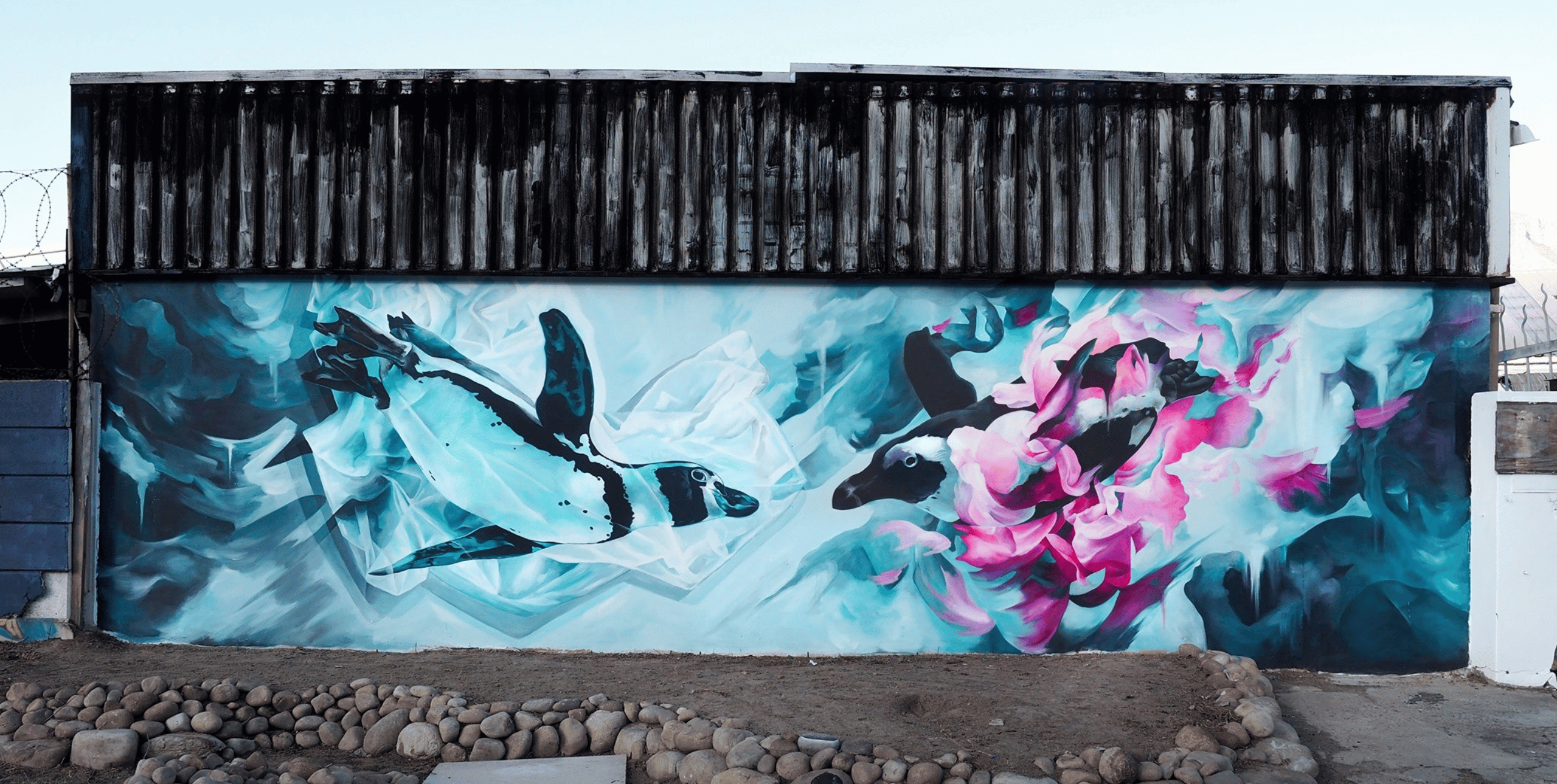 A mural of two penguins swimming underwater on a the wall of a small building with a wooden roof.