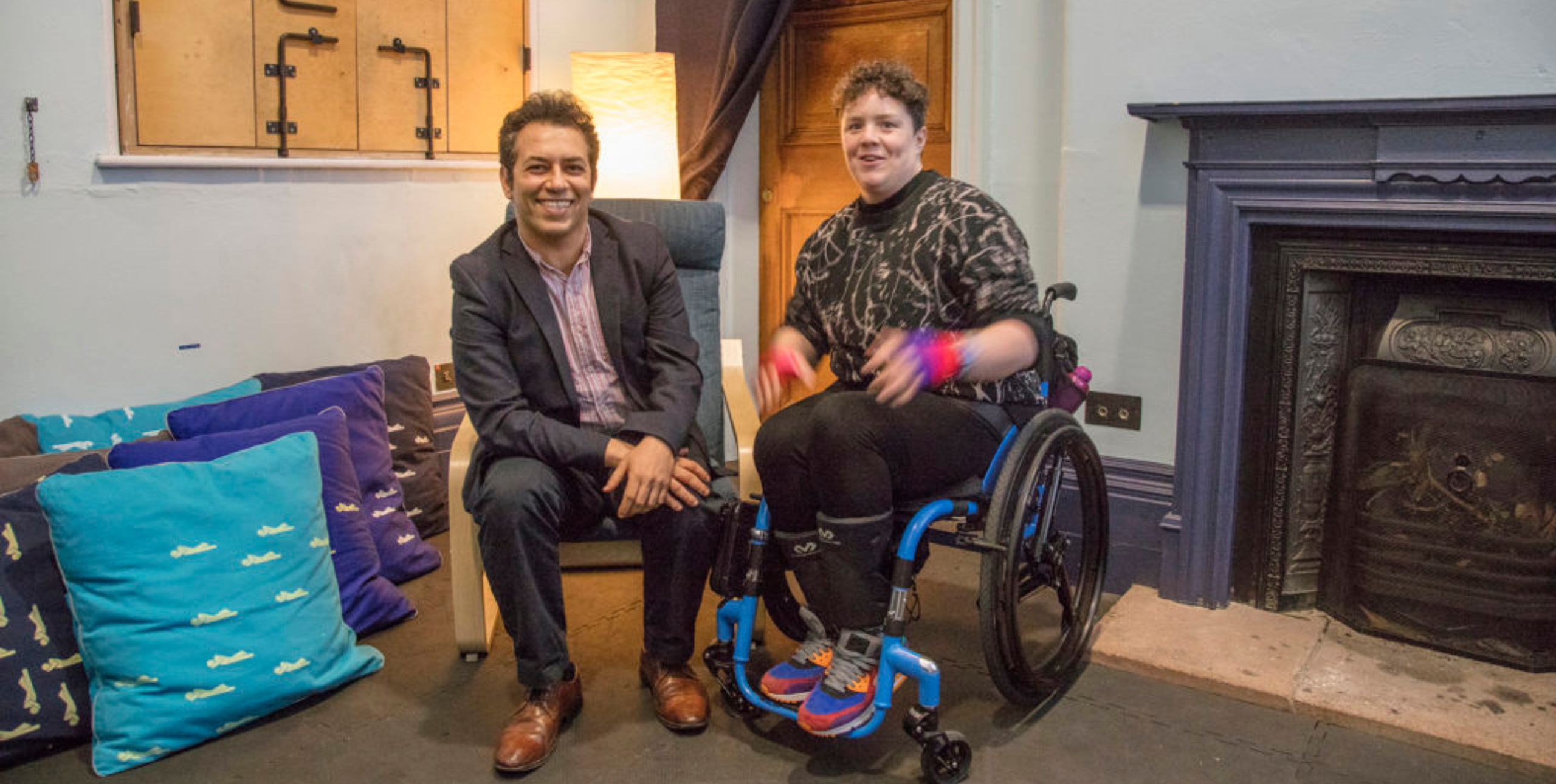 Artistic Director & CEO of Battersea Arts Centre, Tarek Iskander with Jess Thom (Touretteshero) in the new permanent rest space at BAC. They sit alongside each other with a fireplace to their left, and large colourful cushions are piled to their right. Tarek Iskander is seated on a chair, wears a grey suit and brown shoes, and is smiling. Jess Thom is seated in her wheelchair and is wearing a sweatshirt with a graphic print and colourful trainers.
